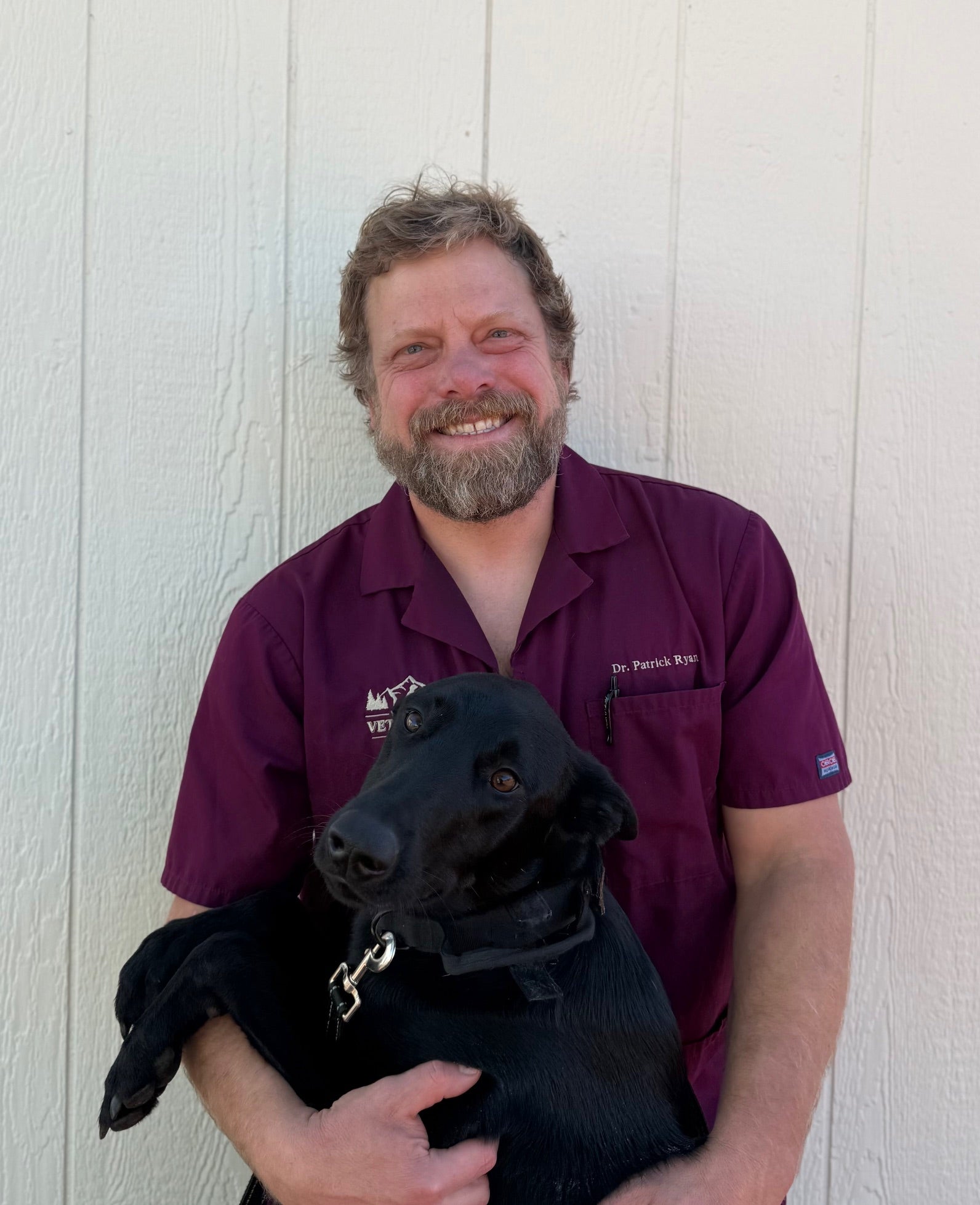 Dr. Ryan wearing a maroon shirt holding his black Belgian Malinois Mix dogog